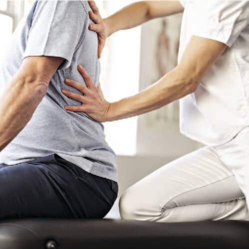 Male patient seated facing outward while physical therapist applies manual therapy techniques on his back to help alleviate pain.